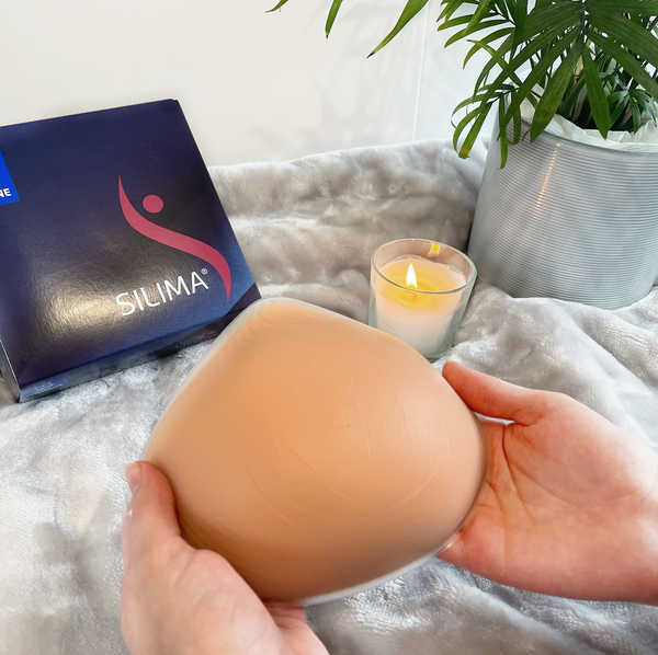 Picture of one Silima Soft & Light Symmetrical Breast Form (Front). In the back of the image is a box with the Silima logo to store the breast form. 