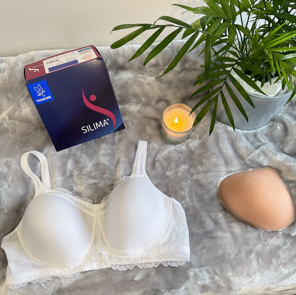Picture of a Silima Soft & Light Symmetrical Breast Form on the right side of the image. To the left is a Silima pocketed bra. In the back of the image, above the bra, is a box with the Silima logo to store the breast form. 